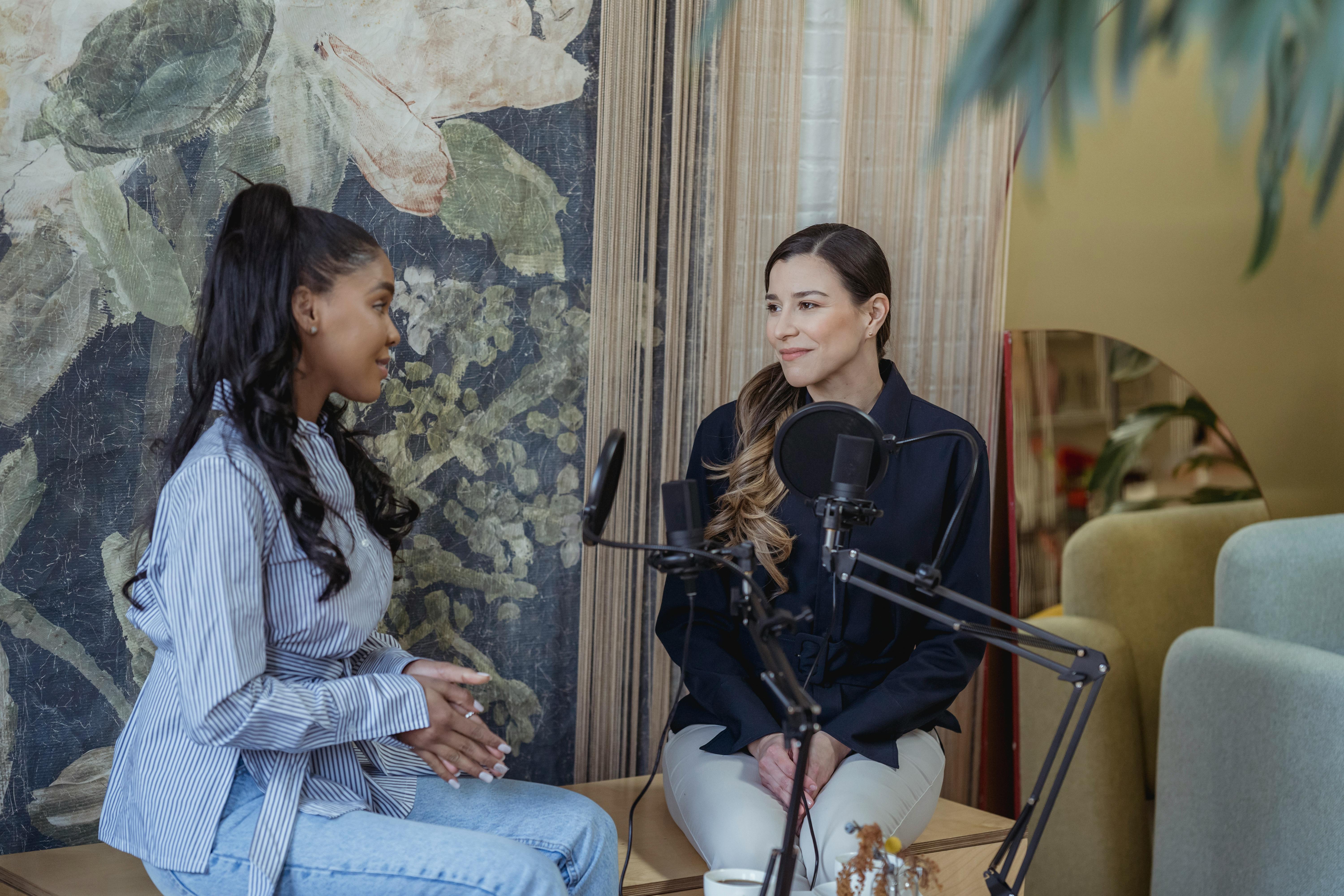 Two women in a cozy setting recording a podcast, conversing with microphones.