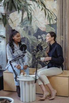 Two women in conversation during a podcast recording, enjoying coffee in a studio setting.