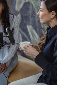 Woman holding a coffee cup speaking into a condenser microphone during a podcast session indoors.