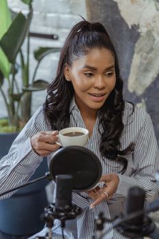 African American woman in striped shirt holding coffee, sitting by microphone indoors.