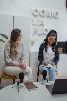 Two women engage in a casual podcast discussion with microphones, notebooks, and a laptop