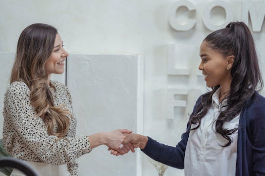 Two professional businesswomen shaking hands in a modern office setting.