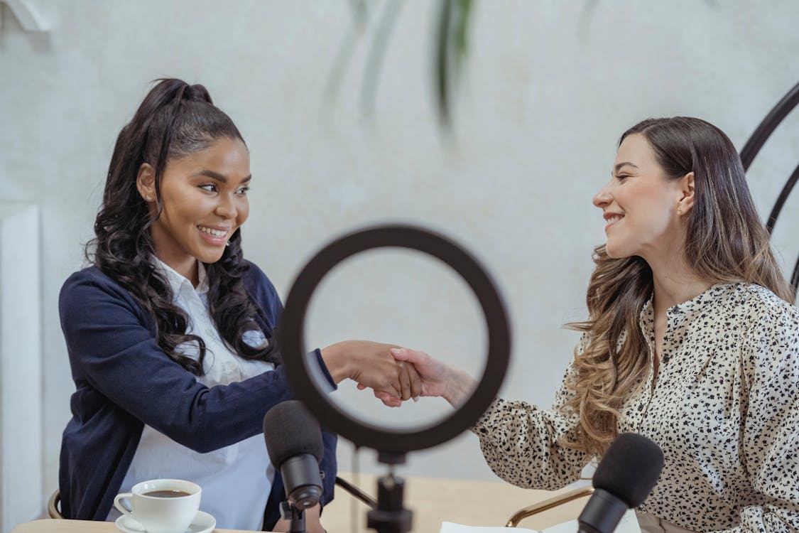 Free Smiling multiethnic females wearing formal outfits shaking hands and looking at each other friendly while sitting against round video light after interview Stock Photo
