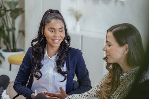 Two women in a friendly conversation at an indoor office setting, one smiling.