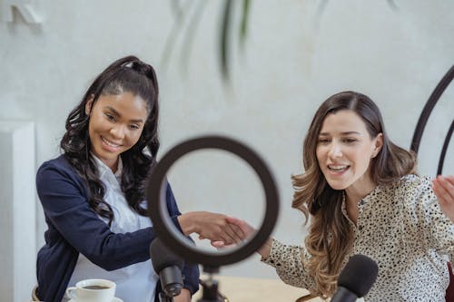 Free Positive young multiracial female colleagues shaking hands and smiling while sitting at table with microphones and coffee cup and recording vlog Stock Photo