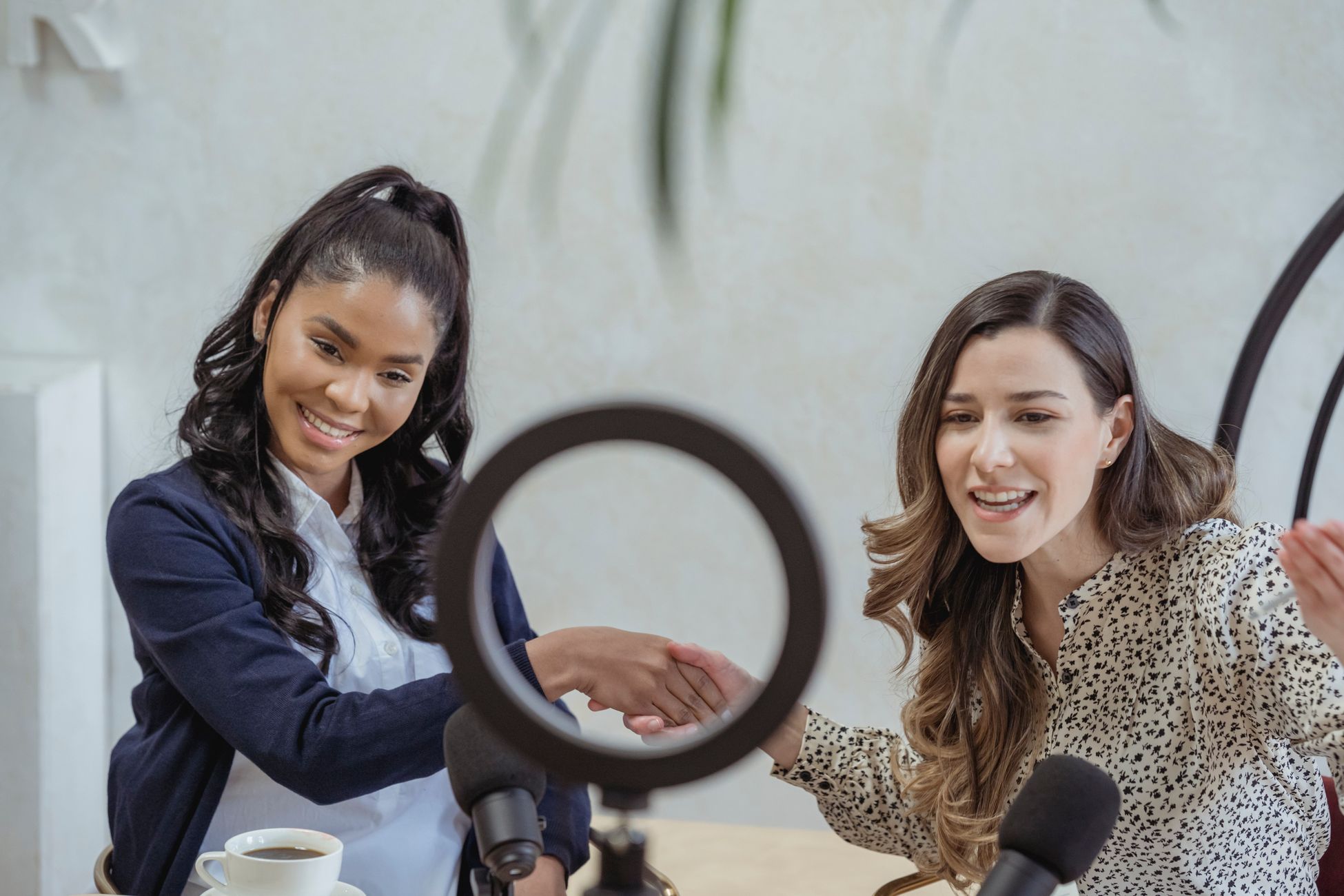 Positive young multiracial female colleagues shaking hands and smiling while sitting at table with microphones and coffee cup and recording vlog