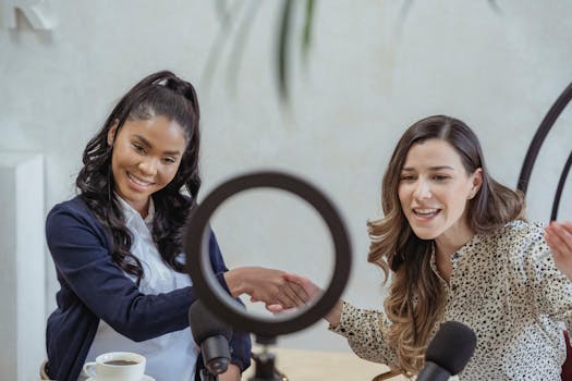 Positive young multiracial female colleagues shaking hands and smiling while sitting at table with microphones and coffee cup and recording vlog