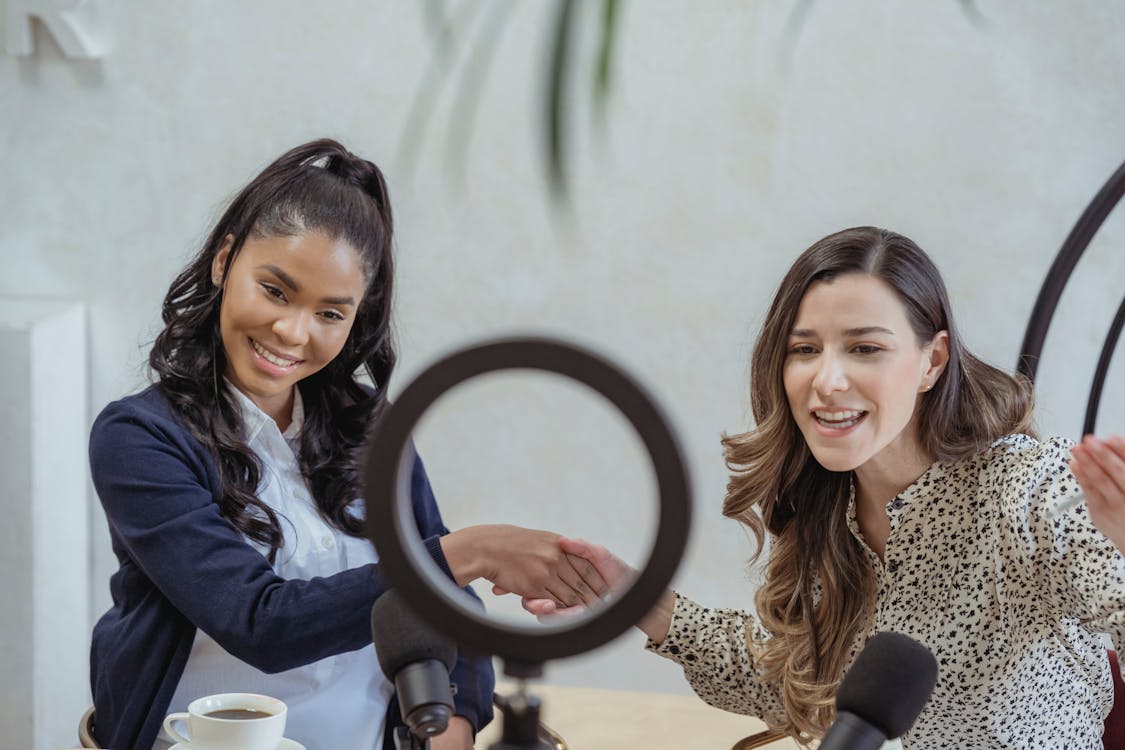 Positive young multiracial female colleagues shaking hands and smiling while sitting at table with microphones and coffee cup and recording vlog