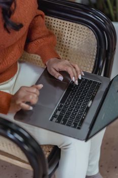 From above of crop anonymous female freelancer in stylish sweater sitting on wicker chair and typing on laptop while working remotely in cafe