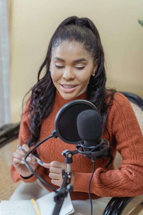 Joyful young African American female radio presenter with notebook on knees in wars sweater talking into microphone during podcast recording in studio