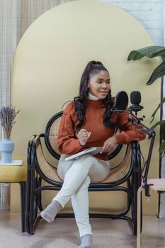 Young black female vlogger with dark long hair in casual outfit sitting on chair with crossed eyes and speaking into microphone while recording podcast at home
