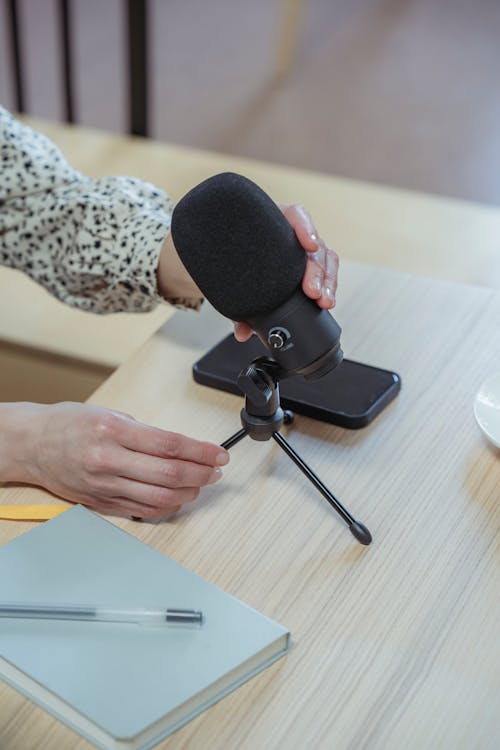 From above of crop unrecognizable female employee setting up microphone while sitting at wooden table with notebook and smartphone during briefing