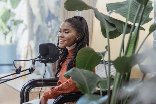 Side view of positive young African American female with long dark hair in casual outfit sitting on chair and singing into microphone while recording podcast in studio