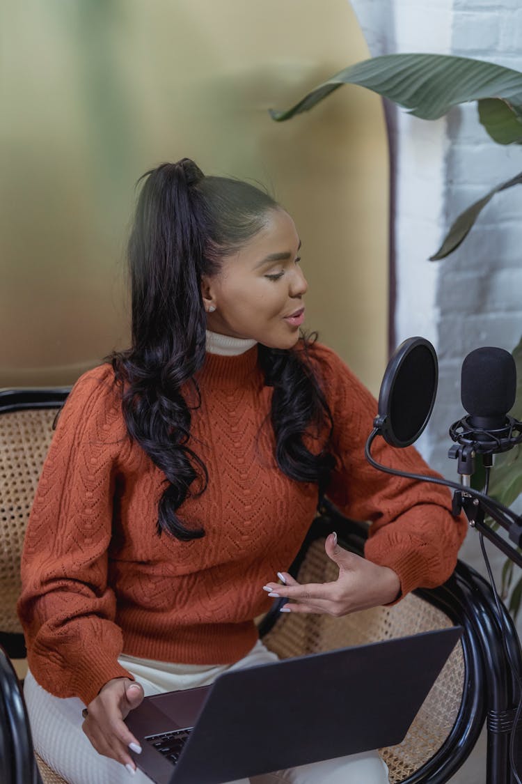 Young Ethnic Woman With Laptop On Knees Recording Podcast At Home