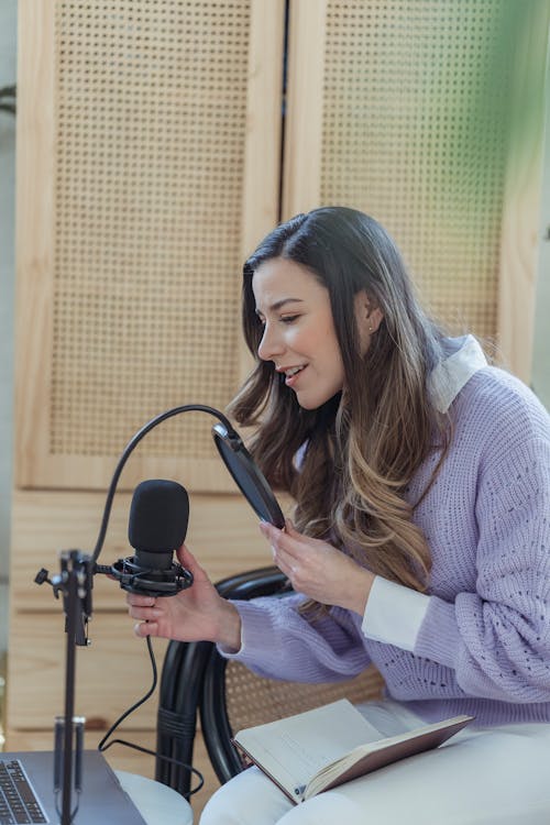 Young positive lady in casual clothes sitting in armchair while recording podcast on microphone with notepad on knees near laptop in light room