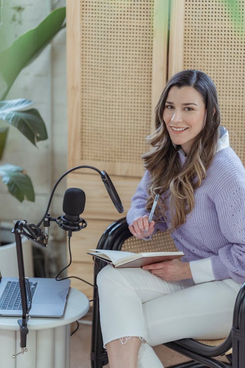 Young positive lady in casual clothes sitting in armchair near laptop and microphone with notepad on knees near cabinet and potted plant while looking at camera in light room