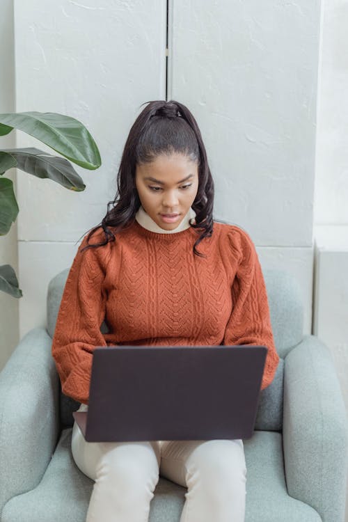 Concentrated African American female teleworker with wavy ponytail sitting in armchair and typing on netbook