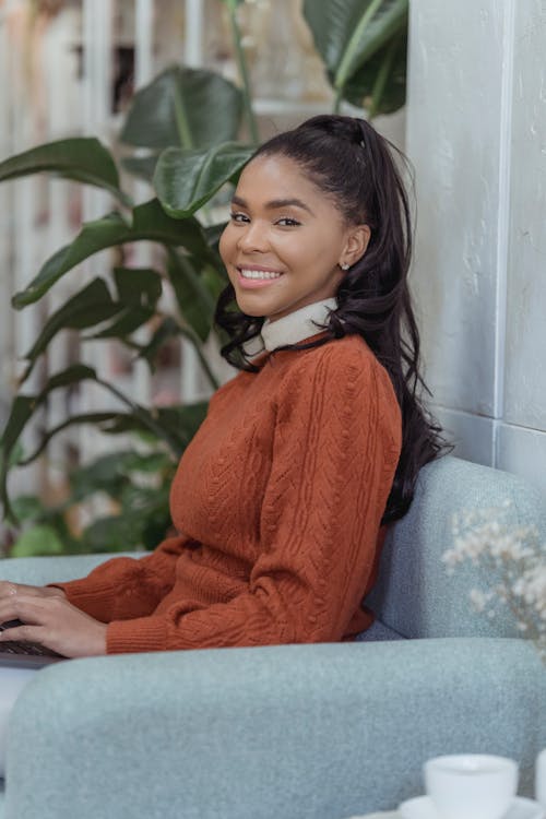 Side view of positive African American female with toothy smile sitting at comfortable armchair and typing on netbook while looking at camera