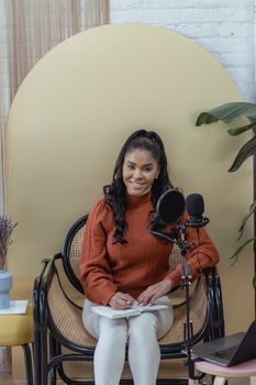 Positive African American female with long wavy hair sitting with notepad and recording audio on mic placed near laptop
