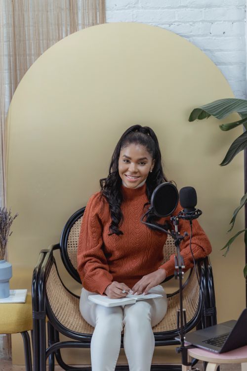 Free Positive African American female with long wavy hair sitting with notepad and recording audio on mic placed near laptop Stock Photo
