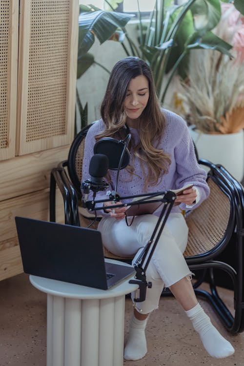 High angle full body if female host sitting in armchair with crossed legs and recording audio on laptop with microphone