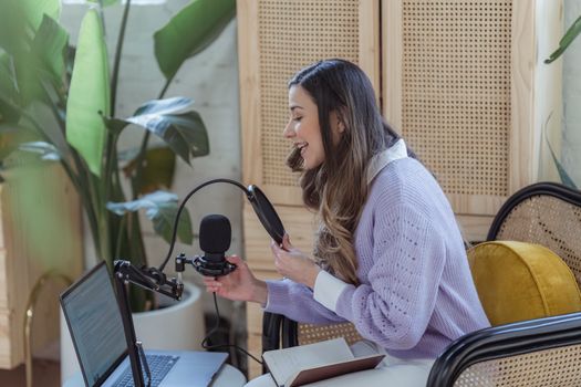 High angle side view of female influencer sitting in armchair and recording podcast on mic connected to laptop