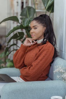 Side view of pensive African American female freelancer sitting in armchair with laptop and thoughtfully looking away