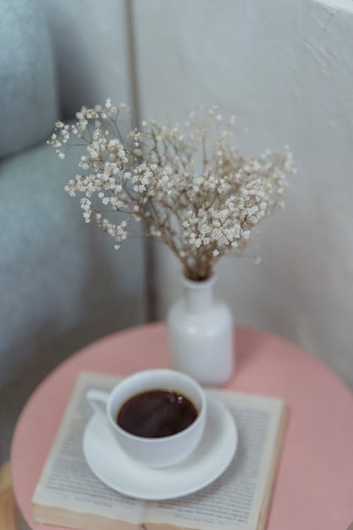 High angle of blooming flowers in ceramic vase placed near cup of aromatic coffee above opened book on table