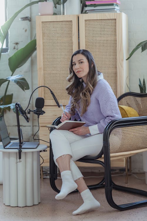 Full body of female sitting in armchair with notepad near laptop and microphone for remote broadcast