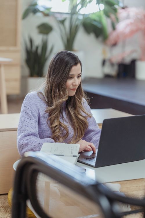 Free Young female freelancer with long hair sitting at table and using netbook for online work sitting at table with opened notebook Stock Photo