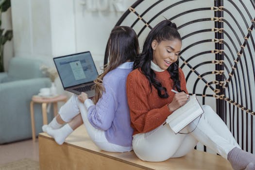 Positive African American female writing in notebook sitting back to back with coworker using netbook