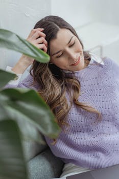 Smiling young woman relaxing on a sofa indoors, engaging with her laptop. Greenery adds a serene touch.