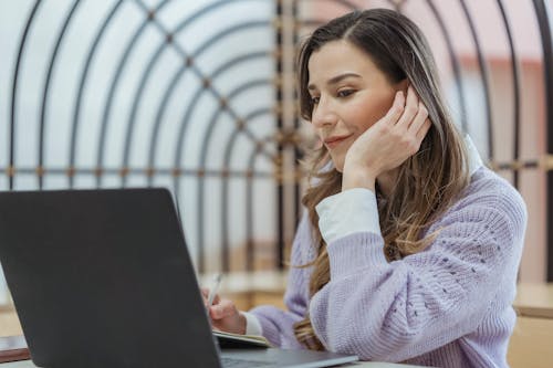 Free Smiling female teleworker leaning on hand while taking notes in planner during remote work at looking at screen Stock Photo
