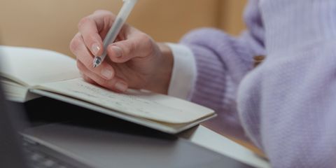 Crop faceless female freelancer working on netbook while writing in planner while sitting at table during remote occupation