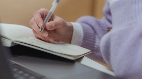 Crop faceless female freelancer working on netbook while writing in planner while sitting at table during remote occupation