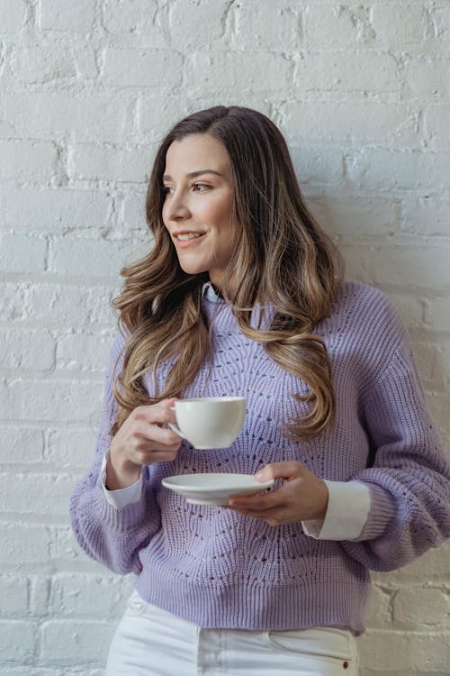 Free Young positive female with long wavy hair drinking hot coffee while leaning on white brick wall Stock Photo