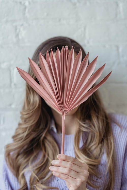 Unrecognizable female with long wavy hair hiding face behind dried sun palm against white brick wall