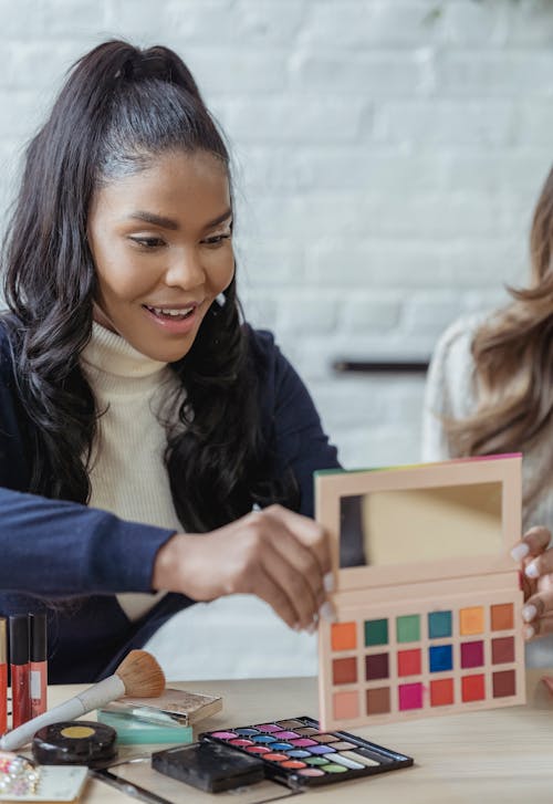 Free Cheerful African American female visagiste showing pallet of eyeshadows while working with makeup products at table with crop colleague Stock Photo