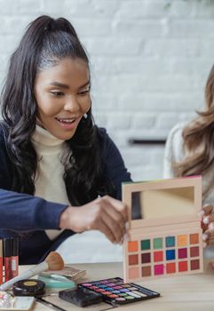 Cheerful African American female visagiste showing pallet of eyeshadows while working with makeup products at table with crop colleague
