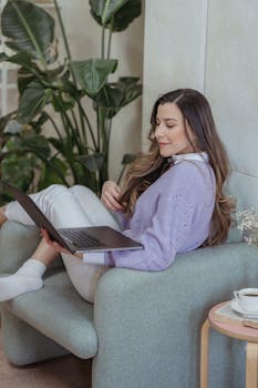 Young woman working on a laptop while lounging in a cozy armchair, embodying modern remote work lifestyle.
