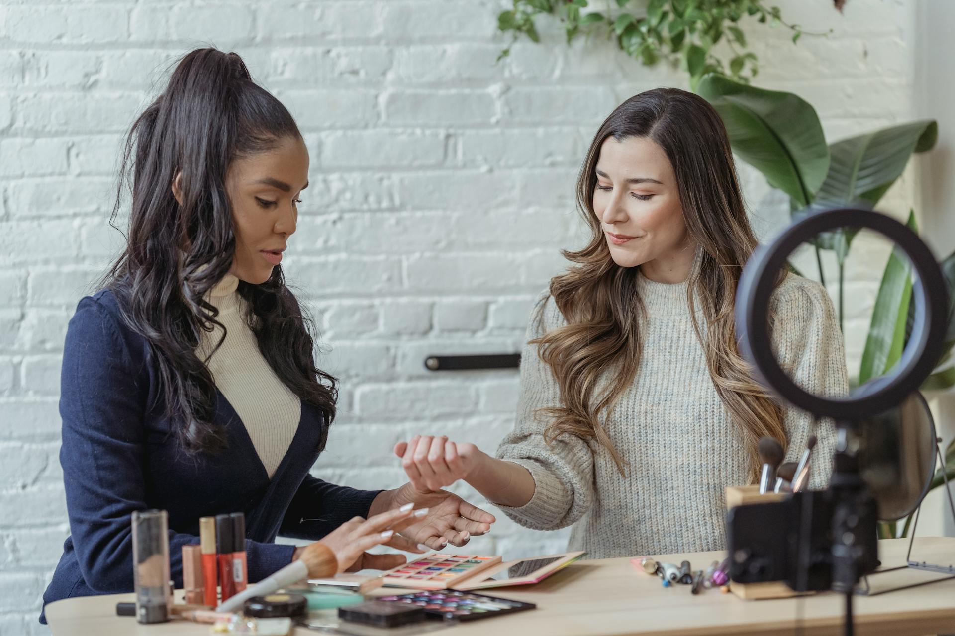 Young positive multiethnic female bloggers testing makeup products while sitting at table and recording vlog on smartphone placed on tripod with ring light