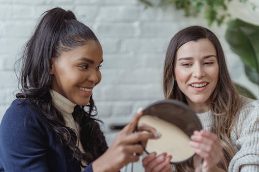 Two women smiling and applying makeup with a mirror indoors.