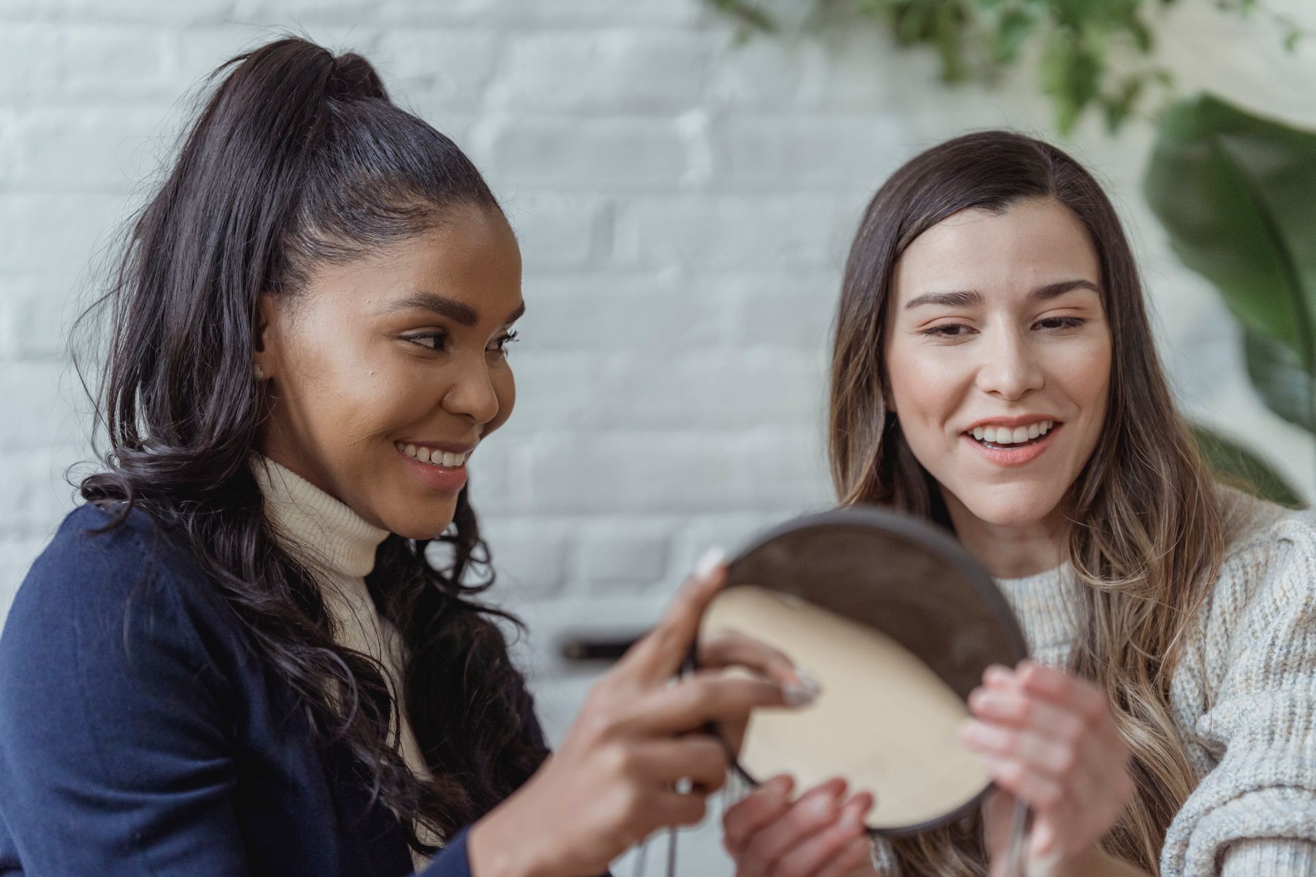 Two women smiling and applying makeup with a mirror indoors.