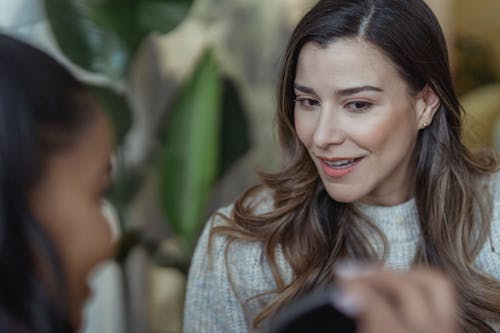 Crop unrecognizable African American female visagiste smiling and holding mirror in front of happy client with long wavy hair in beauty salon