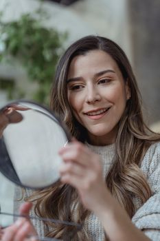Content young female with long brown hair in warm sweater smiling while looking in cosmetic round shaped mirror after applying makeup