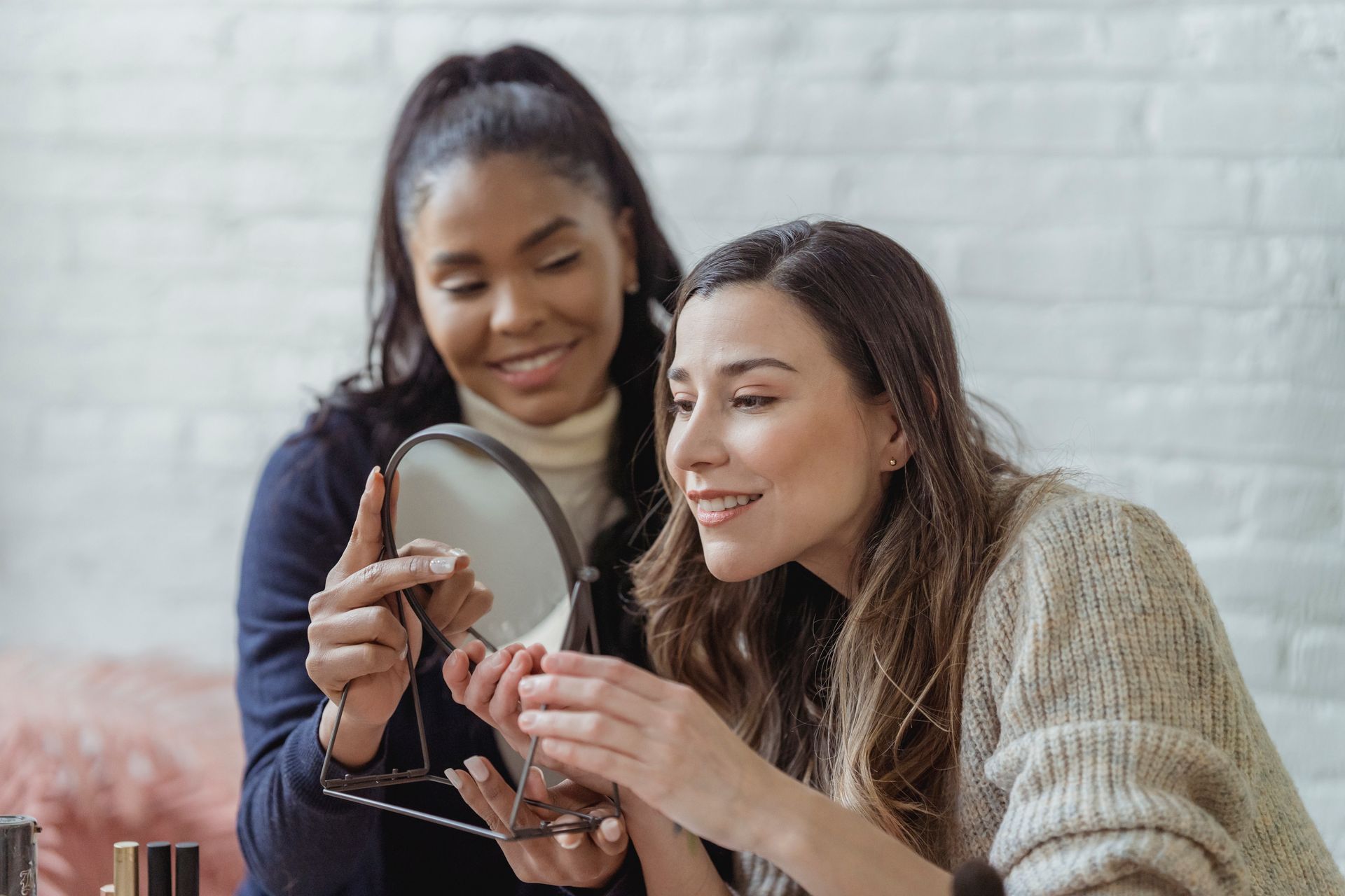 Delighted young woman smiling and looking in round mirror after applying makeup by African American female visagiste in beauty salon