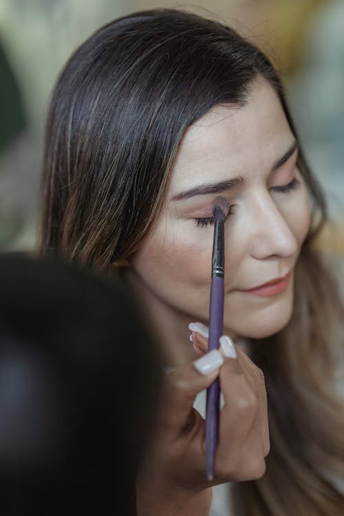 Free From above of crop unrecognizable makeup artist applying eyeshadow with brush on eyelid of young female client sitting in beauty salon with closed eyes Stock Photo