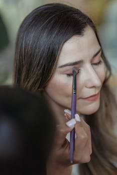 From above of crop unrecognizable makeup artist applying eyeshadow with brush on eyelid of young female client sitting in beauty salon with closed eyes
