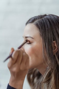 Side view of crop young female customer with ling brown hair and sitting in light studio closed eyes while anonymous makeup artist applying eyeshadows