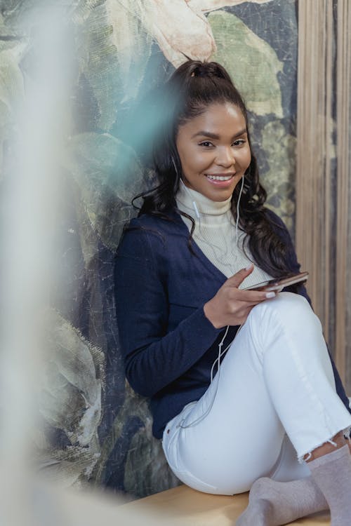 Cheerful young African American female with long dark hair in stylish outfit sitting on floor and home and smiling while using smartphone and listening to songs in earphones